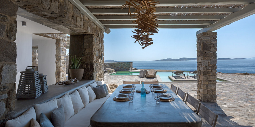 Luxurious villa in Mykonos, with a stunning infinity pool and outdoor lounge area, surrounded by lush greenery. The concierge service is available to assist with all your needs during your stay in this exclusive location.