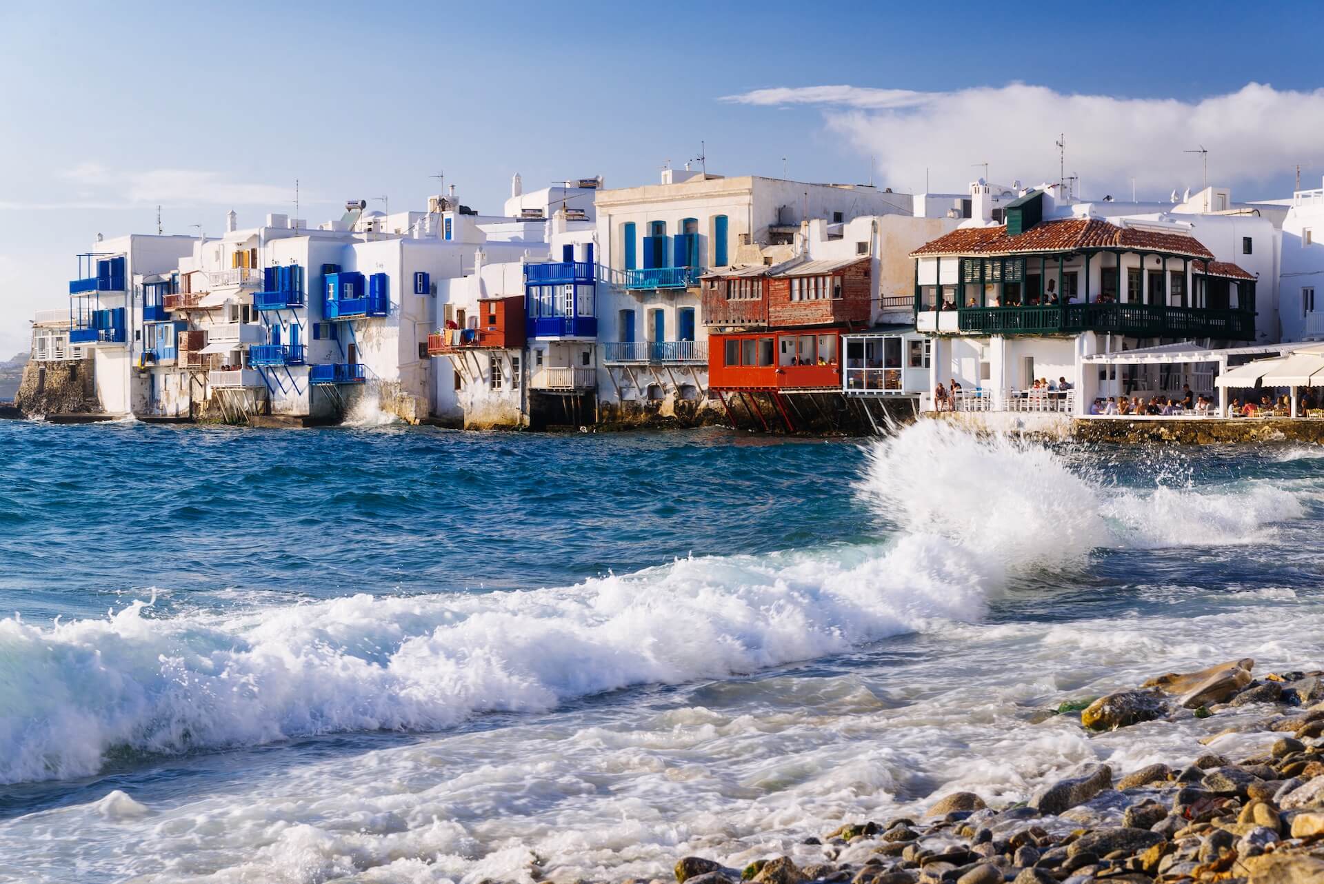 A view of some colorful Mykonian buildings and waves hitting the rocks