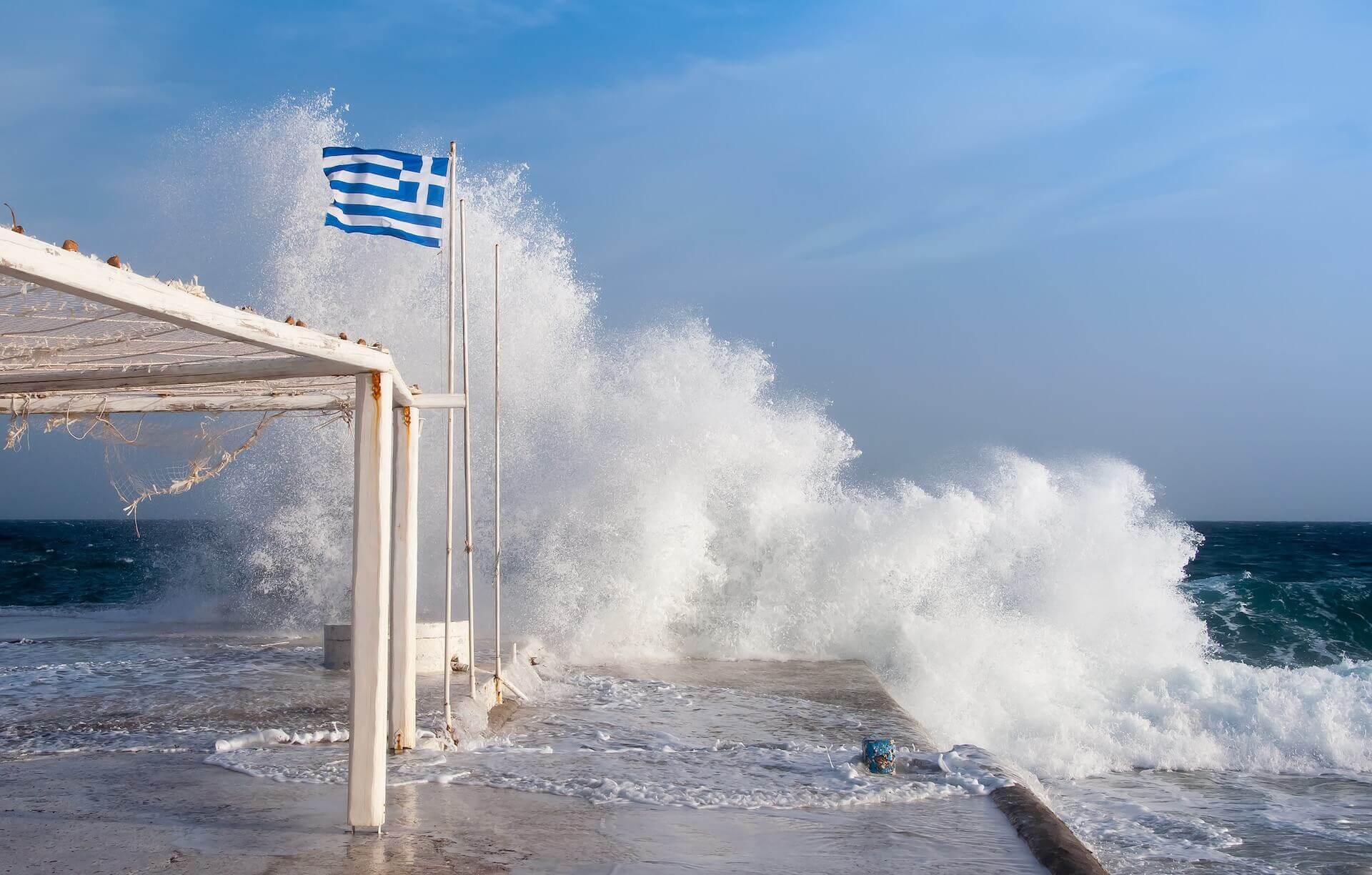 A strong wave hits the dock, where a Greek flag is perched