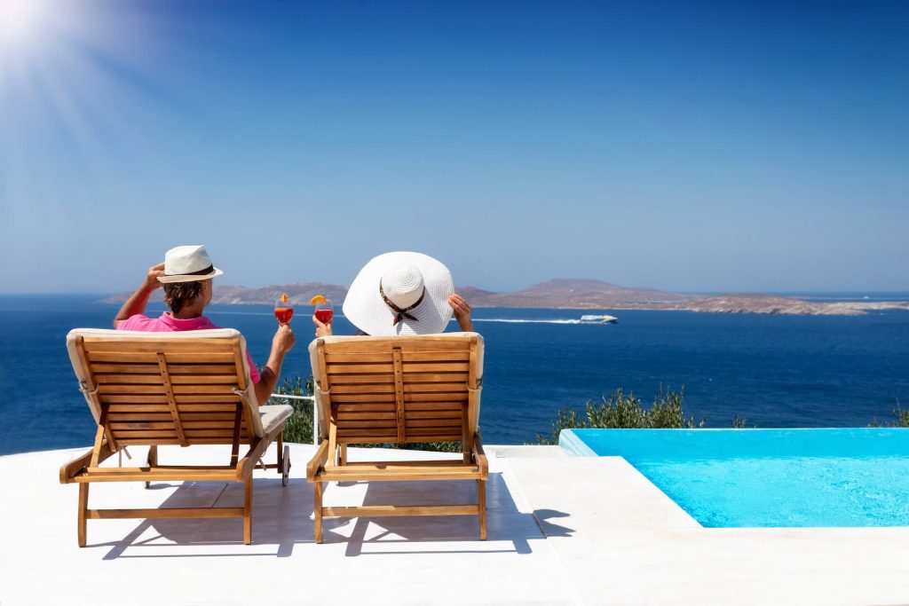 Couple sitting on sunbeds by the pool with cocktails in their hands, facing the sea