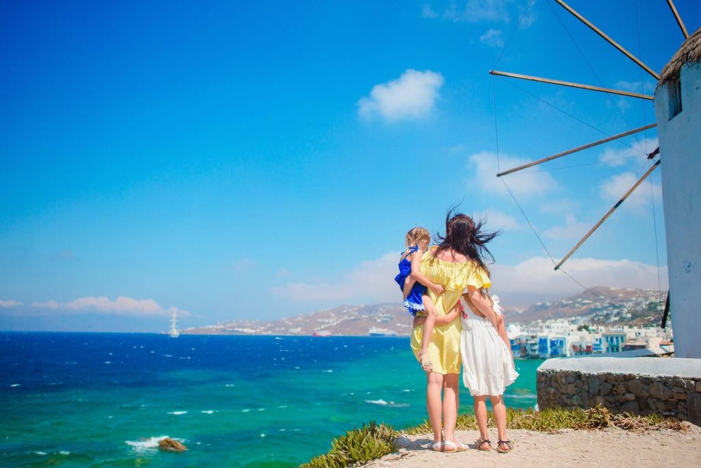 Woman carrying a kid and holding another kid's hand next to a windmill in Mykonos