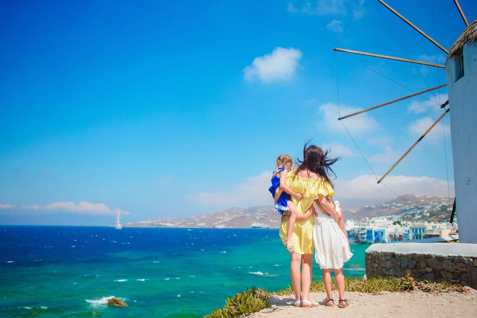 Woman carrying a kid and holding another kid's hand next to a windmill in Mykonos 