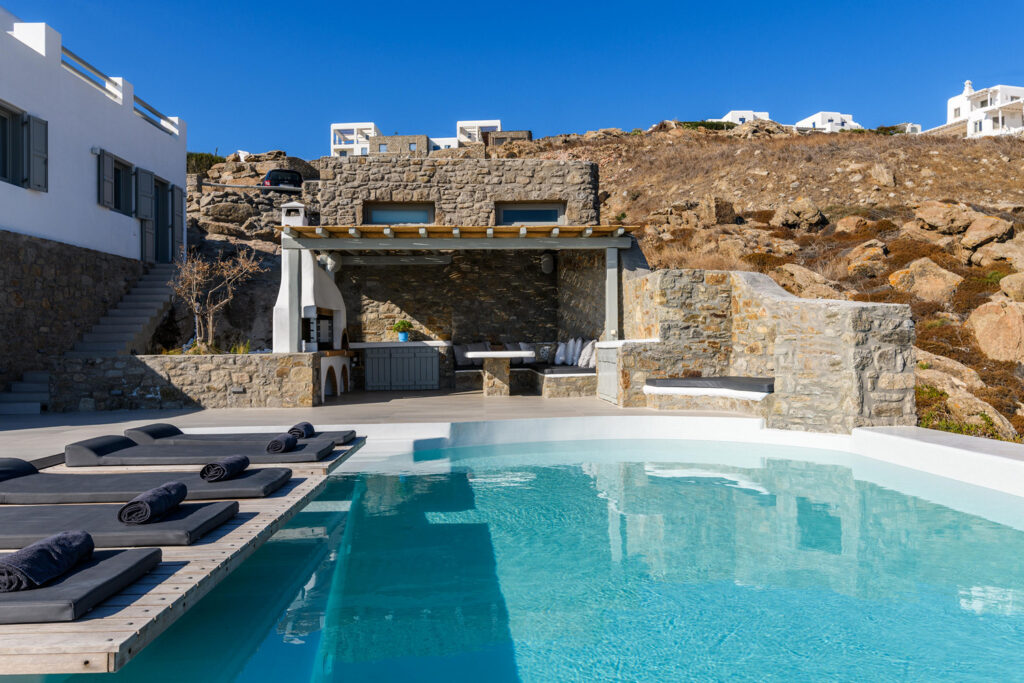 An outdoor area of the Mykonos villa, showcasing a luxurious and spacious pool surrounded by pristine white sun loungers and elegant patio furniture. The crystal clear waters of the pool glisten in the sun, creating a mesmerizing contrast with the stark white surroundings. The overall aesthetic of the area exudes sophistication, comfort, and relaxation, making it the perfect place to spend a lazy day soaking up the sun or enjoying a refreshing swim. The breathtaking views of the surrounding landscape, including the deep blue sea and the distant horizon, serve as the perfect backdrop to this idyllic outdoor oasis.