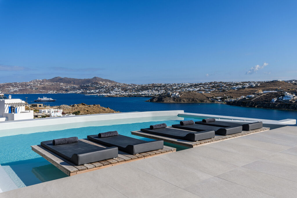 Outdoor lounge area with plush sofas and chairs surrounding a stunning pool overlooking the beautiful scenery of Mykonos.