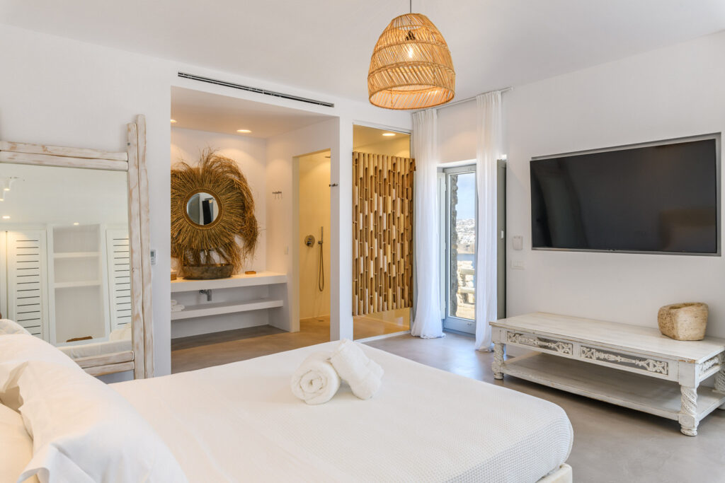A stylish and modern bedroom with white walls, a comfortable looking bed and easy access to a bathroom designed in the traditional Mykonos style, featuring elements such as a distinctive mirror and a luxurious touch that promises to elevate your relaxation experience. The combination of sleek and minimalistic decor with the charming character of Mykonos provides a unique and comfortable atmosphere perfect for a relaxing getaway.