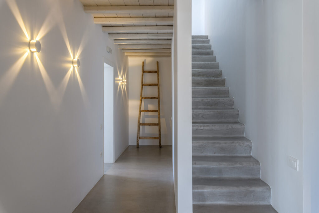 A beautifully lit staircase leading up to the top level of a luxurious villa in Mykonos. The staircase features elegant lights that illuminate the steps and add a warm glow to the space, making it a inviting and relaxing place to be. The attention to detail in the design of the staircase showcases the high-end quality and sophistication of the villa, making it a sought-after destination for discerning travelers.