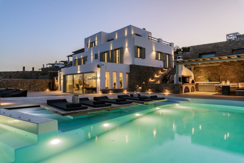 A beautiful outdoor view of the luxurious Mykonos Top Villa, showcasing its lush green garden and inviting infinity pool surrounded by comfortable outdoor seating areas. Enjoy the stunning Mykonos sunset from the comfort of your own private villa with world-class concierge services.