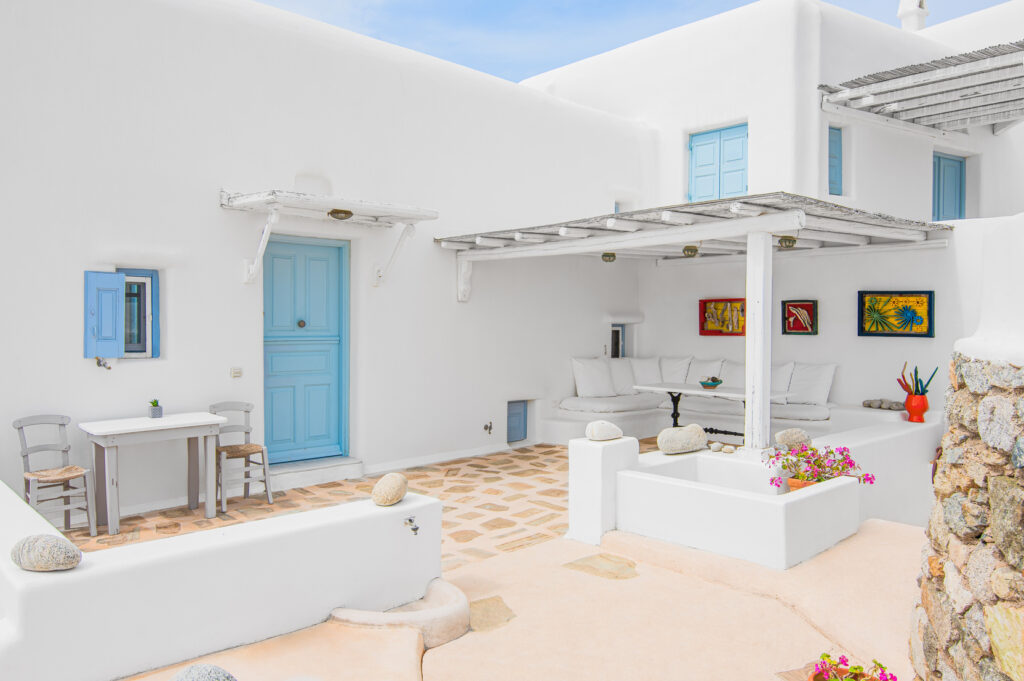 an outdoor lounge area of a luxury villa in Mykonos, with a comfortable seating arrangement surrounded by lush greenery. The space is perfect for relaxing and taking in the stunning surroundings, with the concierge service always on hand to make your stay unforgettable.