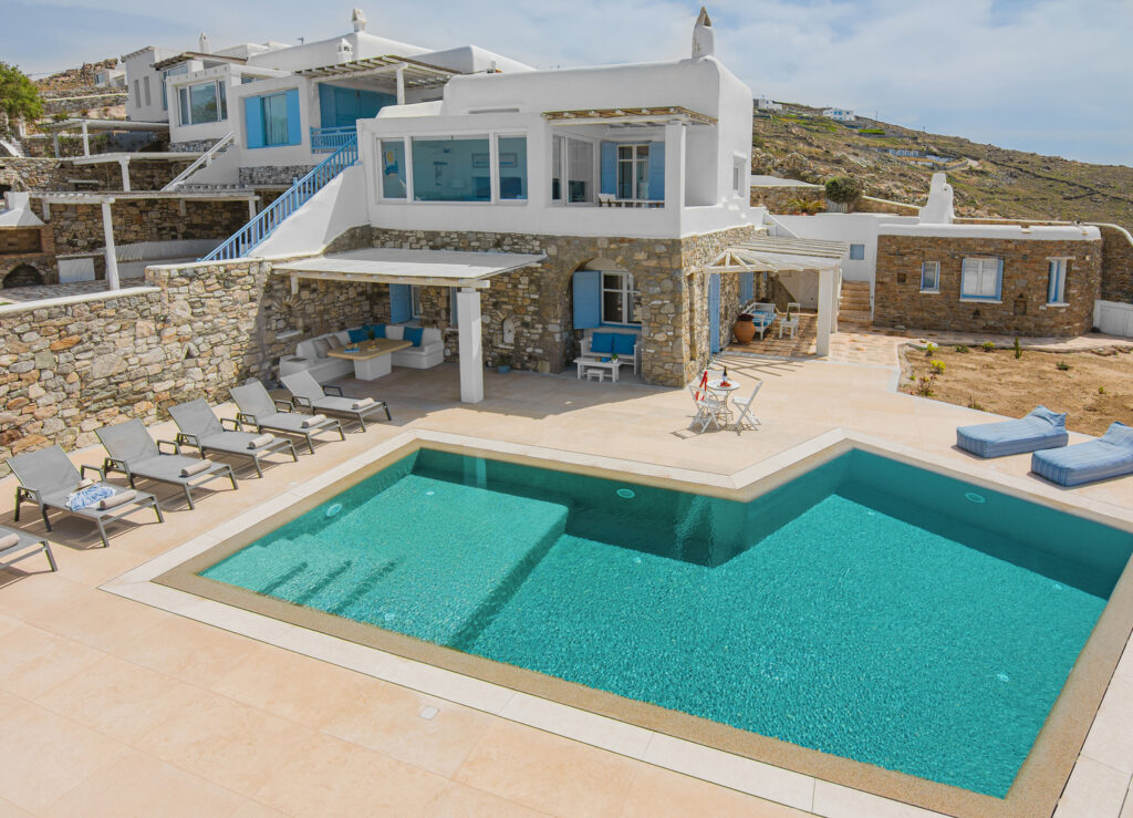 "An outdoor view of a luxurious Mykonos villa with seating and lounging areas, surrounded by greenery and providing a tranquil escape from the bustling city.