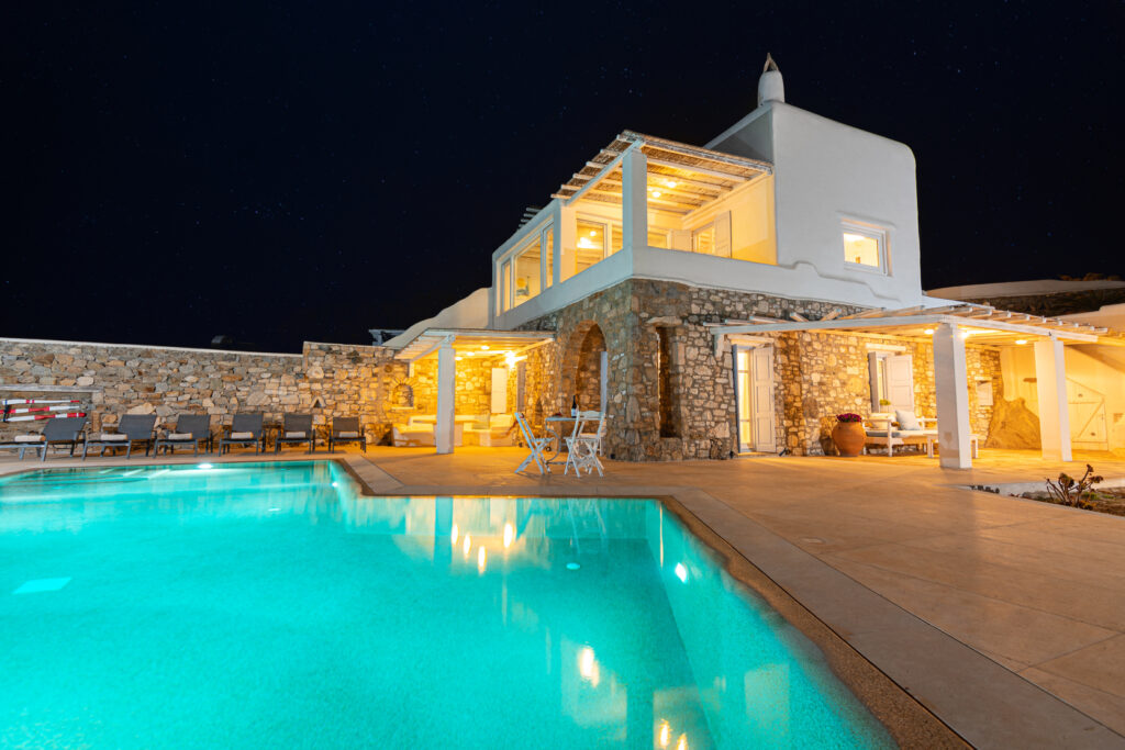 Luxurious outdoor area of a Mykonos villa with a sparkling pool lit up at night. Enjoy concierge service and experience the ultimate in luxury living.