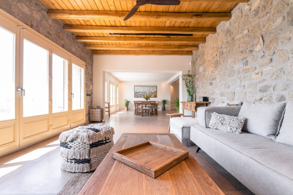 High wooden ceiling, stone walls, and wooden table in the spacious living room, Mykonos rental villa.