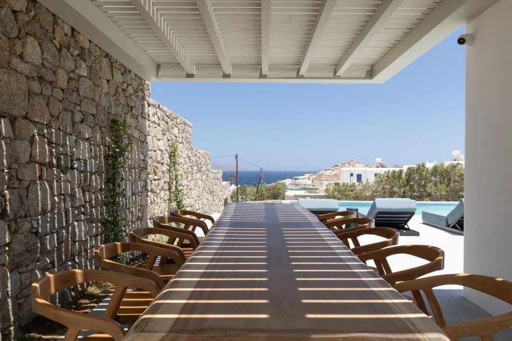 Large wooden dining table in the garden next to private infinity pool of finest rental villa in Mykonos, Greece.