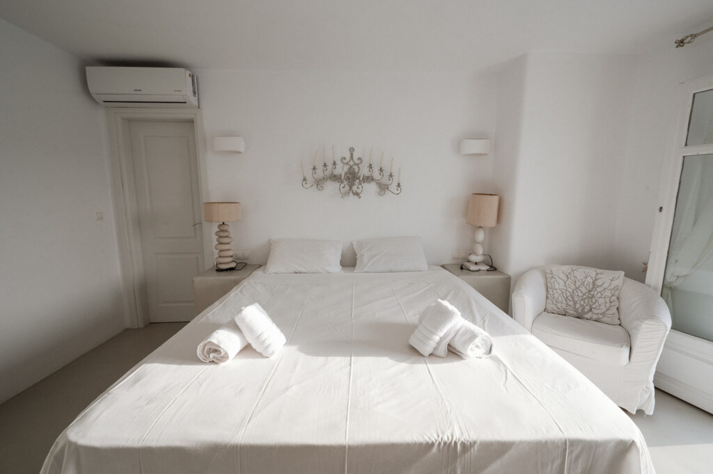 Comfortable and spacious bed, white walls, and an armchair in bedroom in Mykonos splendid villa for booking.