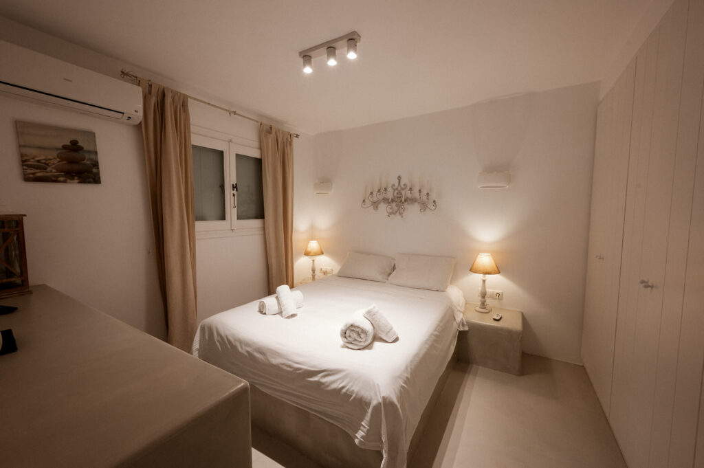 Comfortable stay in a Mykonos villa with a spacious bed, white walls, and soft lighting is available for booking now.
