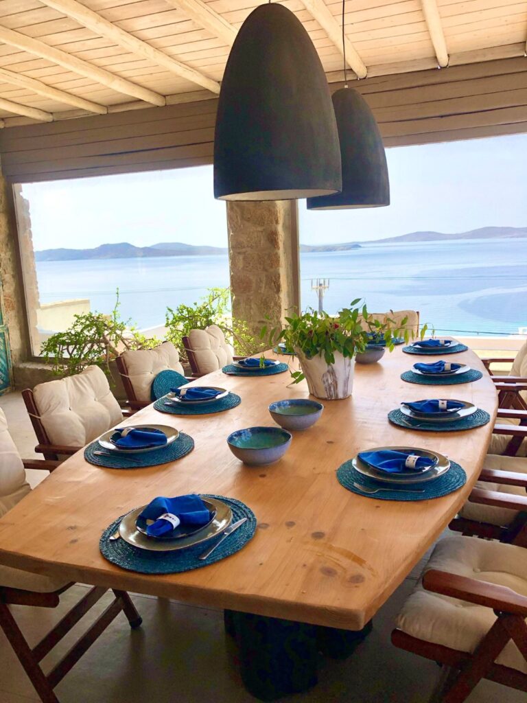The stunning view of the Aegean Sea from the open dining space in Mykonos vacation villa. Title: Experience The Comfort of the most modern Mykonos’ living room