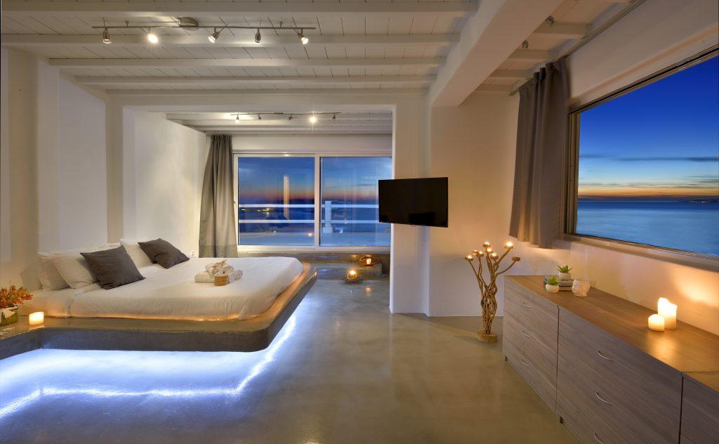 bedroom dimly lit with large windows overlooking the glistening blue sea